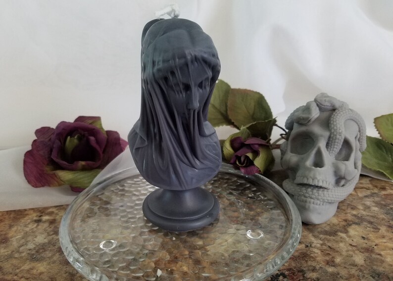 May include: A dark gray candle shaped like a veiled bust, set on a glass plate. A gray skull with snakes and a burgundy rose are also visible. The candle bust has a draped veil over the face.