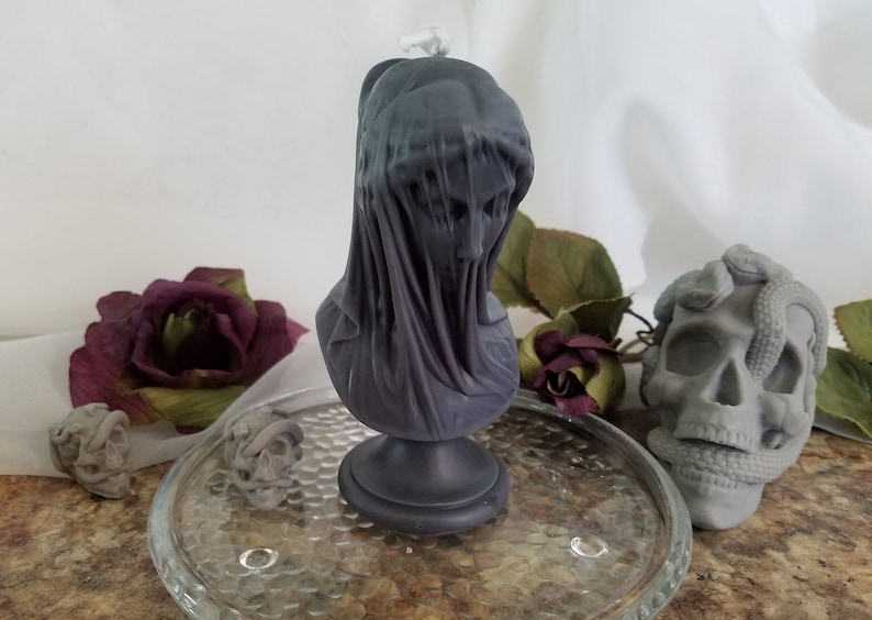 May include: A dark gray candle shaped like a veiled bust, set on a clear glass plate. The candle has a detailed face and draped fabric. Other decorative items include a skull with a snake and a few dark red roses.