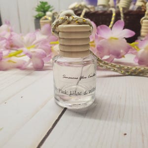 May include: A clear glass bottle with a wooden cap and a rope hanger. The bottle has the text "Sensuous Glow Candles Pink Lilac & Willow". Pink and purple flowers are in the background.