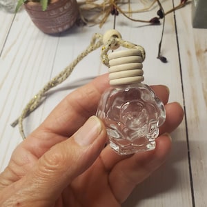 May include: A small, clear glass bottle shaped like a rose, featuring a light-colored, ribbed wooden cap and a braided cord for hanging. The bottle is transparent and designed to hold liquid, with a distinct floral relief pattern on its exterior surface.