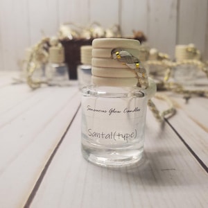 May include: A clear glass bottle with a wooden cap and a decorative cord. The bottle has a label that reads "Sensuous Glow Candles" and "Santal (type)". The bottle is filled with a clear liquid. Other bottles are visible in the background.
