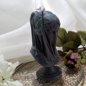 May include: A dark gray candle shaped like a veiled bust. The candle features a detailed sculpture of a face covered by draped fabric. The candle sits on a round base and has a wick at the top. The candle is a decorative item.