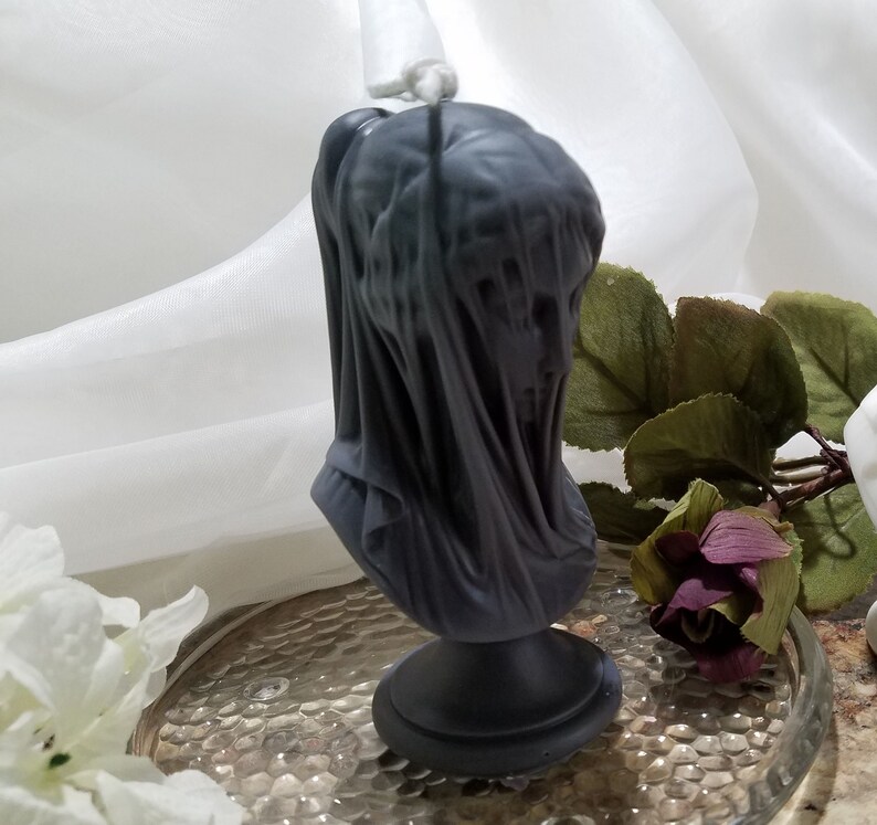 May include: A dark gray candle shaped like a veiled bust. The candle features a detailed sculpture of a face covered by draped fabric. The candle sits on a round base and has a wick at the top. The candle is a decorative item.