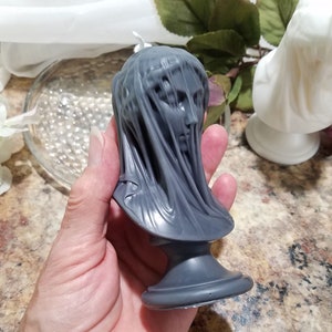 May include: A gray candle shaped like a veiled bust. The candle features a detailed face and draped fabric, resting on a pedestal base. The candle is held in a hand, with a white candle and flowers in the background.
