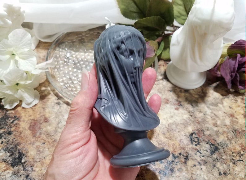 May include: A gray candle shaped like a veiled bust. The candle features a detailed face and draped fabric, resting on a pedestal base. The candle is held in a hand, with a white candle and flowers in the background.