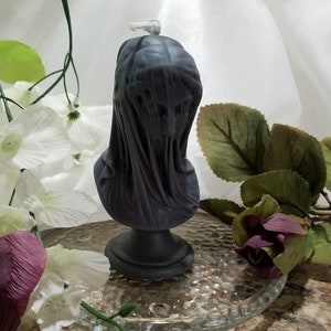 May include: A dark gray candle shaped like a veiled bust, standing on a round base. The candle is surrounded by white and purple artificial flowers and greenery, set on a clear glass plate.