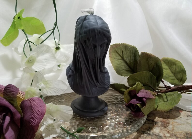 May include: A dark gray candle shaped like a veiled bust, standing on a round base. The candle is surrounded by white and purple artificial flowers and greenery, set on a clear glass plate.