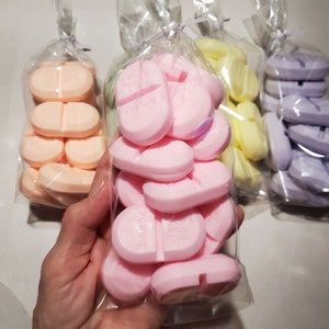 May include: Several bags of colorful, pill-shaped soaps. The soaps are in shades of pink, peach, yellow, and lavender. Each bag contains several soaps, and the soaps have a line down the middle. The soaps are packaged in clear cellophane bags.