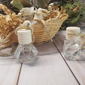 May include: Clear, faceted glass bottles with wooden tops and decorative rope. The bottles are displayed near a woven basket filled with decorative filler and greenery. The bottles are likely for essential oils or perfumes.
