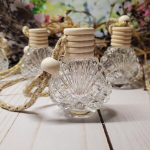 May include: A clear glass bottle with a faceted, shell-like pattern and a ribbed wooden cap. The bottle is suspended by a thick, braided beige cord featuring metallic gold threading and a small, round wooden bead accent for hanging purposes.