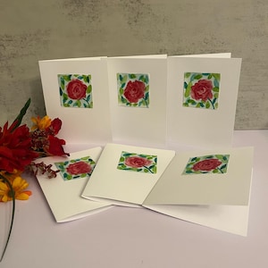 May include: A collection of white greeting cards featuring a watercolor painting of a red rose with green leaves. The cards are arranged in a display, with some standing upright and others fanned out. A bouquet of red and yellow flowers is in the background.