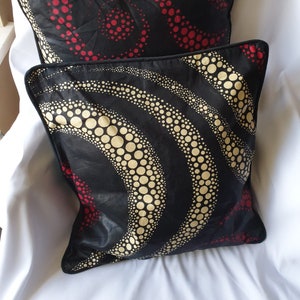 Può includere: Due cuscini decorativi in ​​tessuto nero con un motivo di cerchi rossi e dorati. I cuscini sono su una superficie bianca.