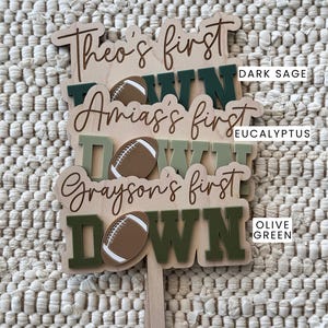 May include: Three wooden signs with the text "Theo's first down", "Amias's first down", and "Grayson's first down". Each sign features a brown football and is painted in a different shade of green: dark sage, eucalyptus, and olive green.