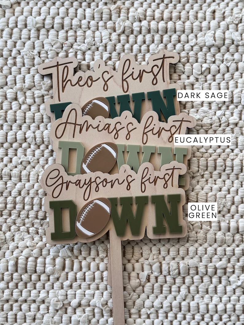 May include: Three wooden signs with the text "Theo's first down", "Amias's first down", and "Grayson's first down". Each sign features a brown football and is painted in a different shade of green: dark sage, eucalyptus, and olive green.
