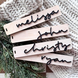 May include: Four wooden gift tags with names written in black script. The names are Melissa, John, Christy, and Ryan. The tags are on a white knitted surface.