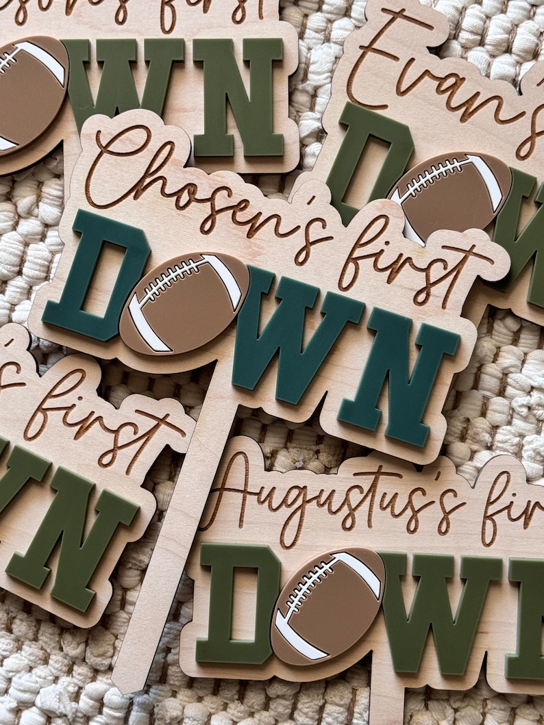 May include: Three wooden signs with the words "Chosen's first down", "Evan's first down", and "Augustus's first down" written in cursive. Each sign has a brown football with white laces and a green outline.