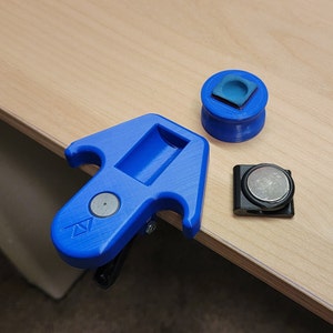Puede incluir: Una herramienta azul impresa en 3D con un clip, un objeto redondo azul y negro con un pequeño cuadrado en la parte superior y un objeto negro con un imán redondo plateado.