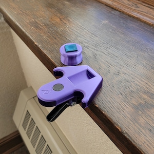 May include: A purple plastic window lock with a black lever and a blue square button. The lock is on a wooden surface.