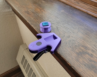 Puede incluir: Una cerradura de ventana de plástico morado con una palanca negra y un botón cuadrado azul. La cerradura está sobre una superficie de madera.