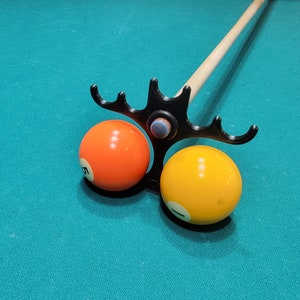 Puede incluir: Un porta-tacos de billar negro con dos bolas de billard, una naranja y una amarilla, unidas al porta-tacos. El porta-tacos está unido a un taco de billar de madera.