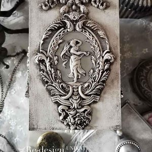 May include: A white wooden box with a decorative silver design featuring a woman in a flowing gown. The box has a distressed finish and is adorned with intricate details. The text "Re-design UNIKAT KAROLINA KARBOWSKA" is visible on the bottom of the box.