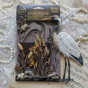 May include: A gray and white crane figurine stands next to a silicone mold. The mold features a design of tall grass, flowers, and a crane. The mold is labeled "DECOR MOULDS" and measures 5" x 8".