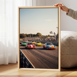 May include: A framed vertical wall art print depicting a vintage car race on an asphalt track. Several colorful sports cars, including red, yellow, green, and blue-orange models, are driving toward a finish line where a person waves a checkered flag.