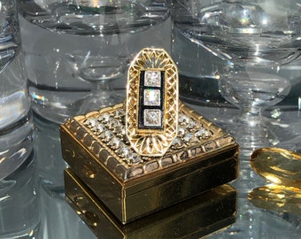 Può includere: Un anello d'oro con una fascia in smalto nero e tre pietre quadrate trasparenti. L'anello è appoggiato su una scatola dorata con un motivo a diamante.