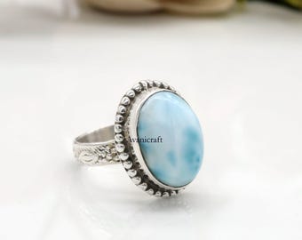 May include: A silver ring featuring a large, oval-shaped, light blue gemstone with white marbling. The gemstone is set in a beaded bezel. The ring band has a floral design. The text "Avanicraft" is visible on the gemstone.