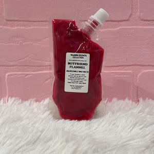 May include: A red, squeezable wax melt pouch with a white spout. The label reads "Boyfriend Flannel" from the Yajaira Scents Collection. The pouch is filled with a deep red wax and sits on a white, fluffy surface.