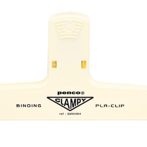 Könnte beinhalten: Ein cremeweißer Kunststoff-Binderclip in T-Form mit einer texturierten Grifffläche an der Oberseite. Auf der Vorderseite ist der Text "penco", "CLAMPY", "BINDING PLA-CLIP" und "ref : 3255464" über einer dreieckigen Grafik aufgedruckt.