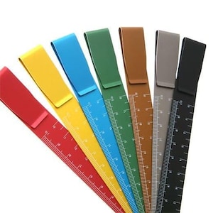 May include: A set of seven metal rulers in red, yellow, blue, green, brown, grey, and black. Each ruler features a clip at the top and white measurement markings along the side, displaying increments in centimetres and millimetres.