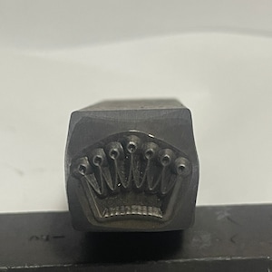 May include: A close-up of a metal stamp with a crown design. The crown has five points and is surrounded by a circle of small dots.