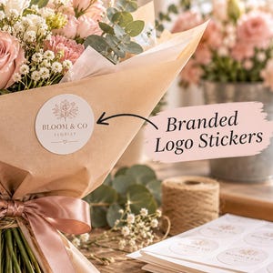 Könnte beinhalten: Ein Blumenstrauß aus rosa und weißen Rosen, eingewickelt in braunes Papier mit einem runden weißen Aufkleber, der den Text BLOOM & CO FLORIST und eine botanische Illustration zeigt. Ein Satinband ist um die Stiele gebunden, daneben liegt ein Bogen Aufkleber.