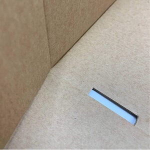 May include: A light brown cardboard box interior featuring a smooth, matte surface with a visible corner seam where two panels meet. A single, rectangular slot is cut into the base panel, revealing a glimpse of light through the opening.