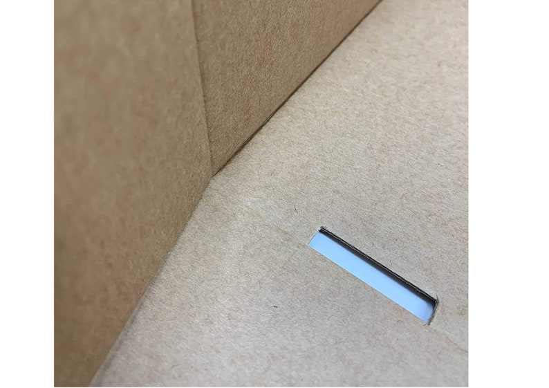 May include: A light brown cardboard box interior featuring a smooth, matte surface with a visible corner seam where two panels meet. A single, rectangular slot is cut into the base panel, revealing a glimpse of light through the opening.