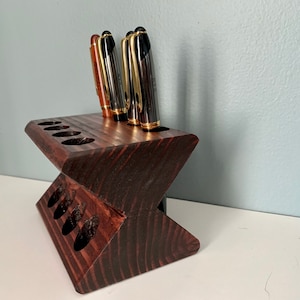 May include: A dark brown wooden pen holder with eight slots for pens. The holder is shaped like a V and has a dark wood grain.
