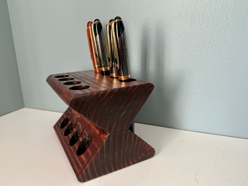 May include: A dark brown wooden pen holder with eight slots for pens. The holder is shaped like a V and has a dark wood grain.