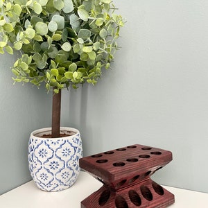 Puede incluir: Un soporte de madera de color morado oscuro con m&uacute;ltiples agujeros para guardar objetos, como pinceles o l&aacute;pices. El soporte tiene forma de reloj de arena y presenta un dise&ntilde;o decorativo. Una planta en maceta con hojas verdes se encuentra en el fondo.