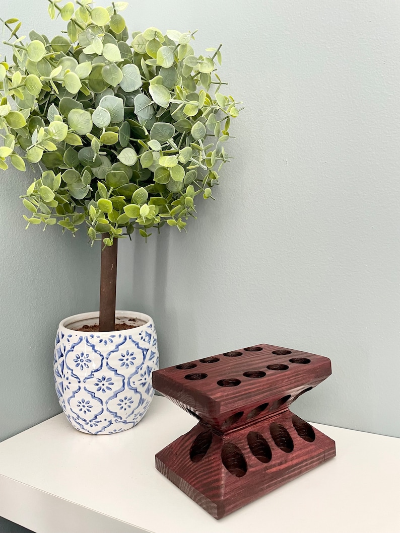 Puede incluir: Un soporte de madera de color morado oscuro con m&uacute;ltiples agujeros para guardar objetos, como pinceles o l&aacute;pices. El soporte tiene forma de reloj de arena y presenta un dise&ntilde;o decorativo. Una planta en maceta con hojas verdes se encuentra en el fondo.