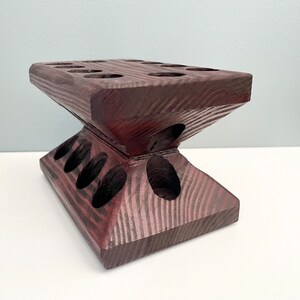 Puede incluir: Un soporte de madera marr&oacute;n oscuro con m&uacute;ltiples agujeros para sostener objetos. El soporte tiene forma de pir&aacute;mide con una parte superior plana.