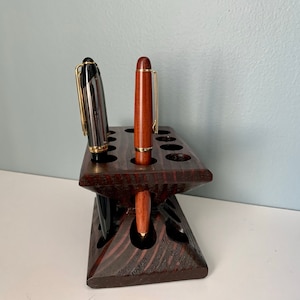 Puede incluir: Un portaplumas de madera marr&oacute;n oscuro con 12 ranuras para plumas. Hay dos plumas en el portaplumas, una negra y otra marr&oacute;n.