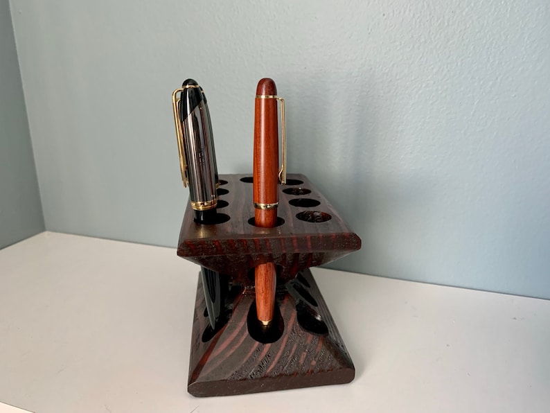 Puede incluir: Un portaplumas de madera marr&oacute;n oscuro con 12 ranuras para plumas. Hay dos plumas en el portaplumas, una negra y otra marr&oacute;n.