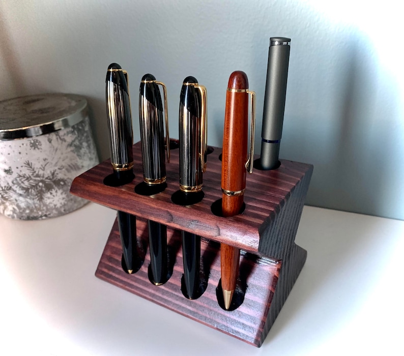 May include: A dark wood pen holder with six slots for pens. Five pens are in the holder, including four black and gold pens and one brown pen. The holder is on a white surface.