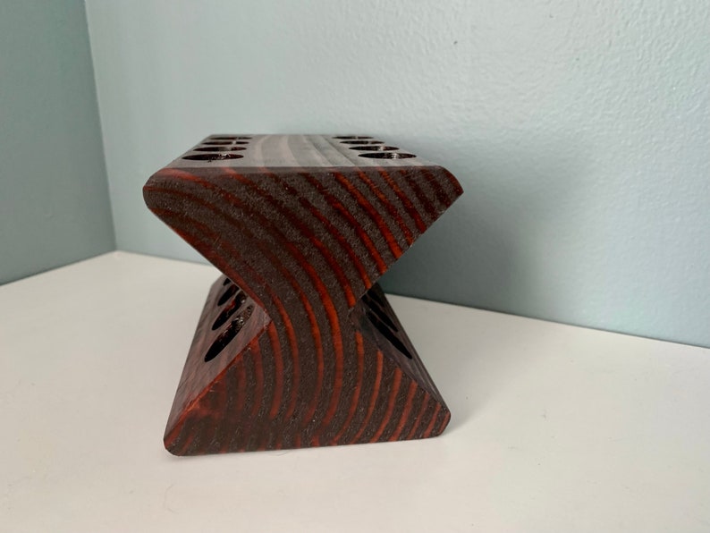 May include: A dark brown wooden stand with multiple slots for holding items, such as pencils or paintbrushes. The stand is shaped like a V and has a smooth, polished finish.