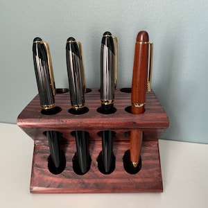 May include: A wooden pen holder with a dark red stain. The holder has eight slots and is angled. Five pens are in the holder, including four black pens with gold accents and one brown pen with a gold accent.