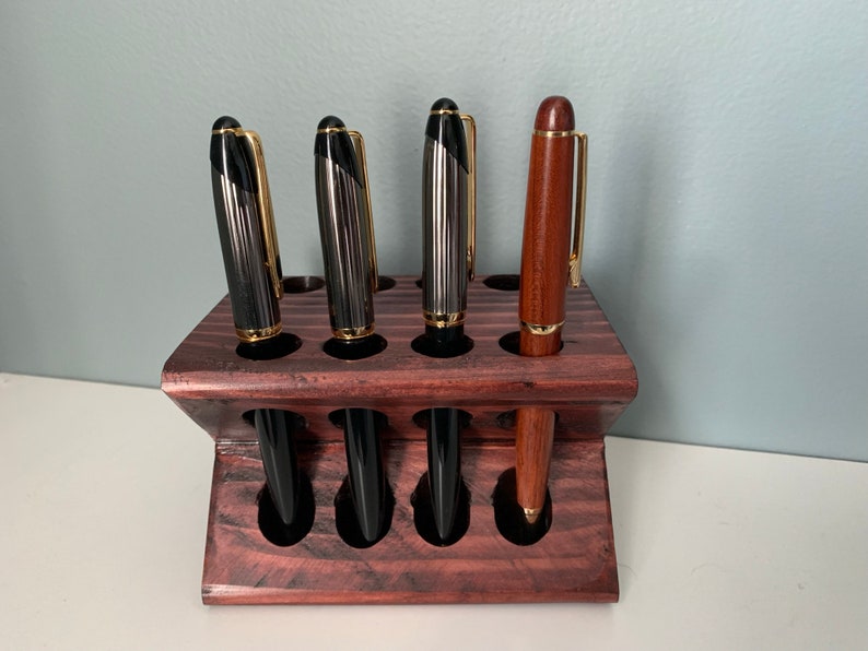 May include: A wooden pen holder with a dark red stain. The holder has eight slots and is angled. Five pens are in the holder, including four black pens with gold accents and one brown pen with a gold accent.