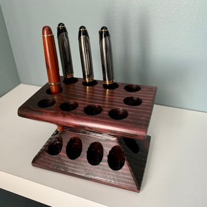 Puede incluir: Un portaplumas de madera marr&oacute;n oscuro con 15 ranuras para plumas. El portaplumas tiene dos niveles y est&aacute; hecho de madera oscura con veta natural. Hay cuatro plumas en el portaplumas, tres negras y una marr&oacute;n.
