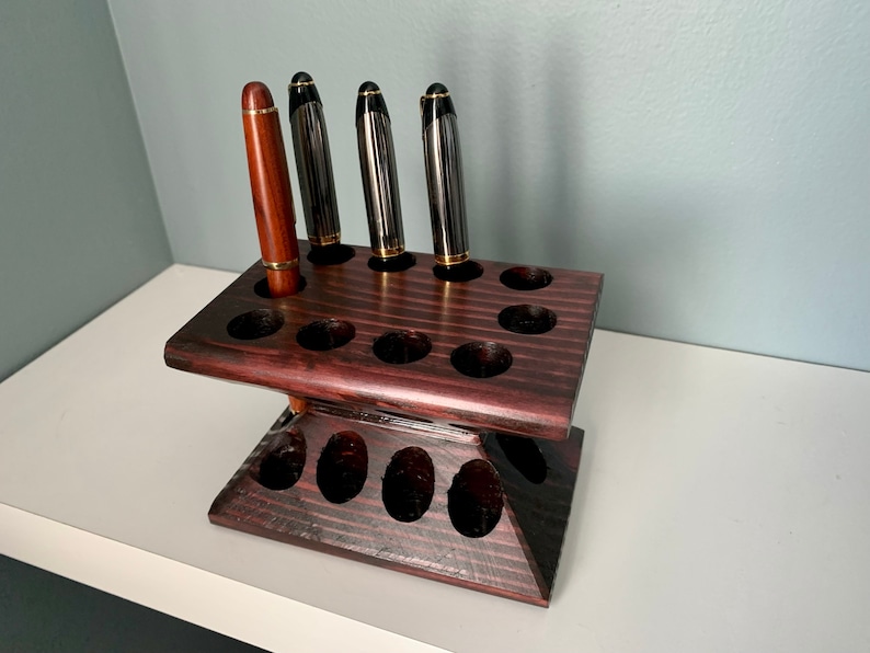Puede incluir: Un portaplumas de madera marr&oacute;n oscuro con 15 ranuras para plumas. El portaplumas tiene dos niveles y est&aacute; hecho de madera oscura con veta natural. Hay cuatro plumas en el portaplumas, tres negras y una marr&oacute;n.