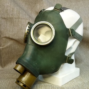 Puede incluir: Una máscara de gas verde con una lente clara y circular y un filtro de color caqui. La máscara está montada sobre una cabeza de maniquí blanca. La máscara tiene correas ajustables y una cubierta facial de goma.